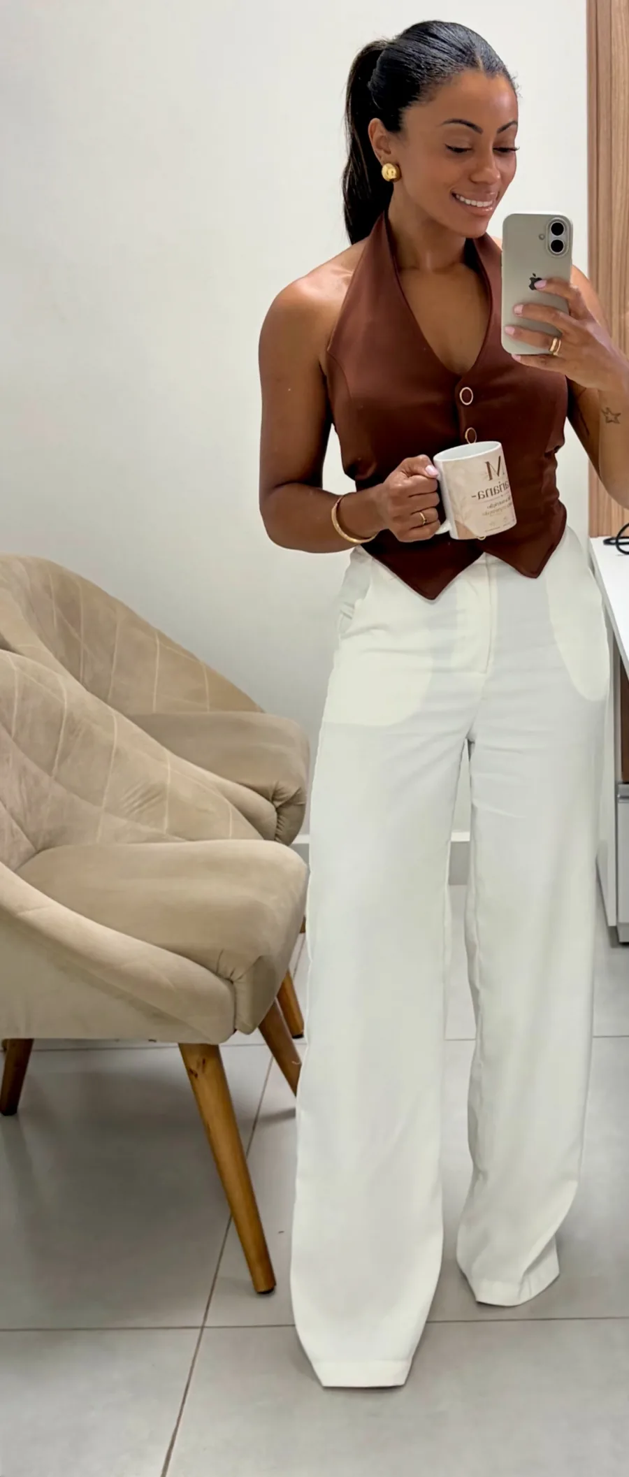 Mariana no consultório, de colete marrom e calça branca, ao lado de uma poltrona